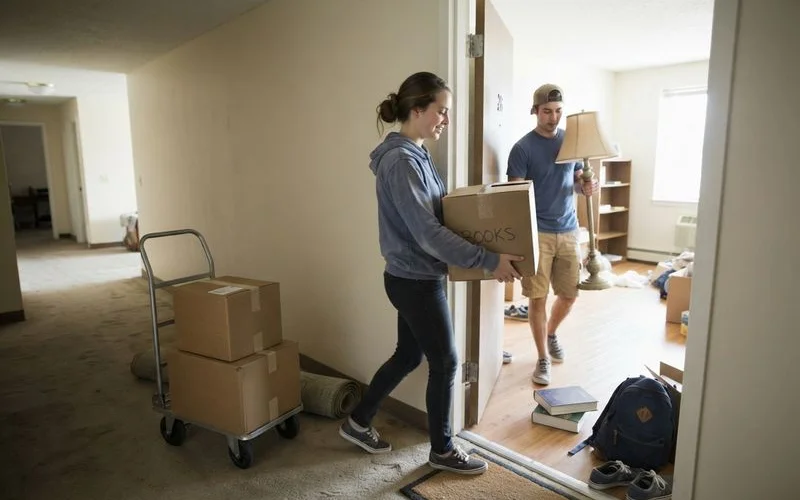Two college students carry boxes and a lamp into a new off-campus apartment near a university
